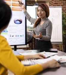 Marketing consultant standing and presenting growth data to coworker seated at office desk. Female business advisor gesturing to device screen, showing her colleague the company analytics summary.